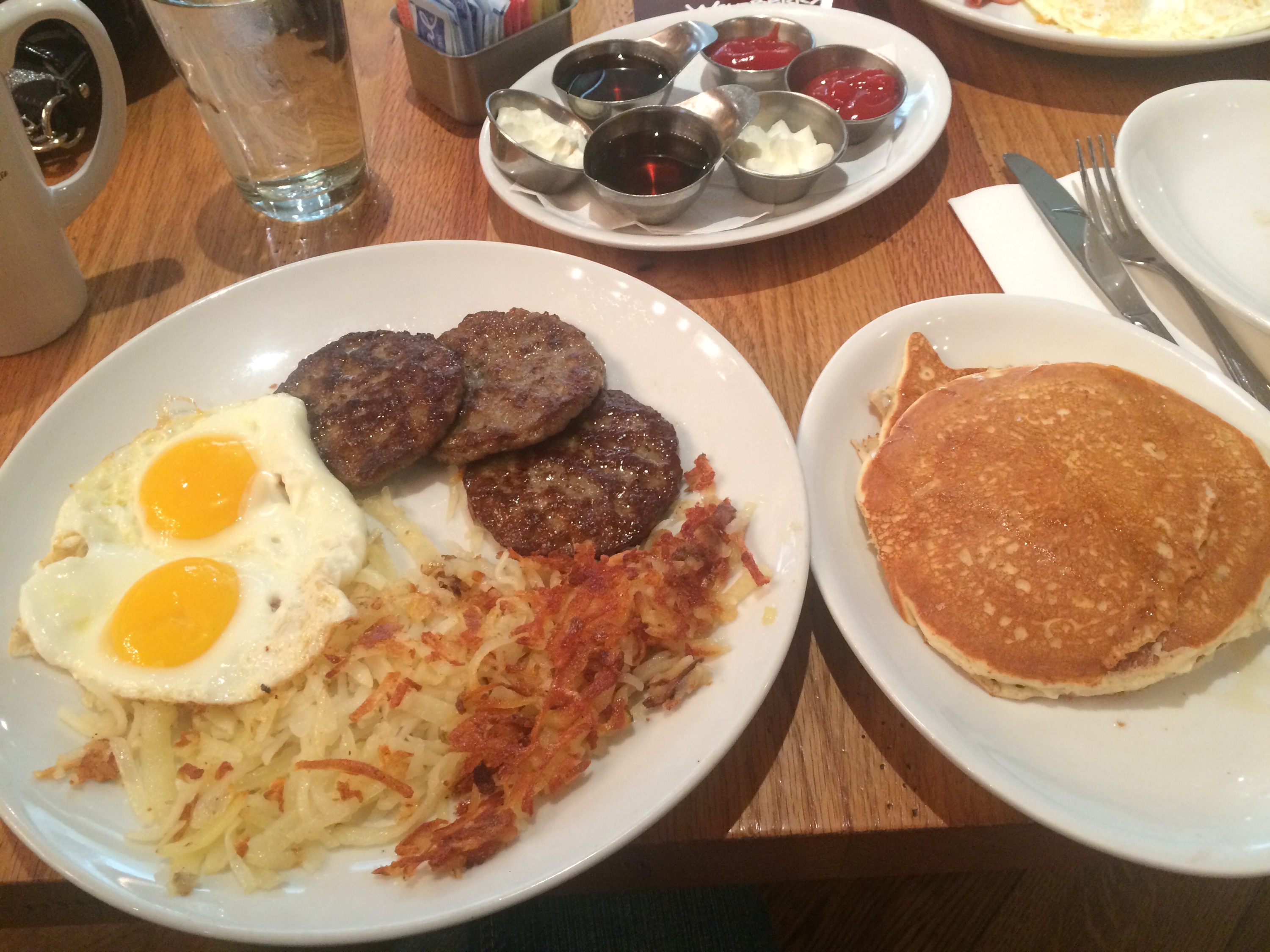 photo of a plate with eggs, hash browns, and sausage patties, and another plate of pancakes. Butter and toppings are on a smaller plate in the background.